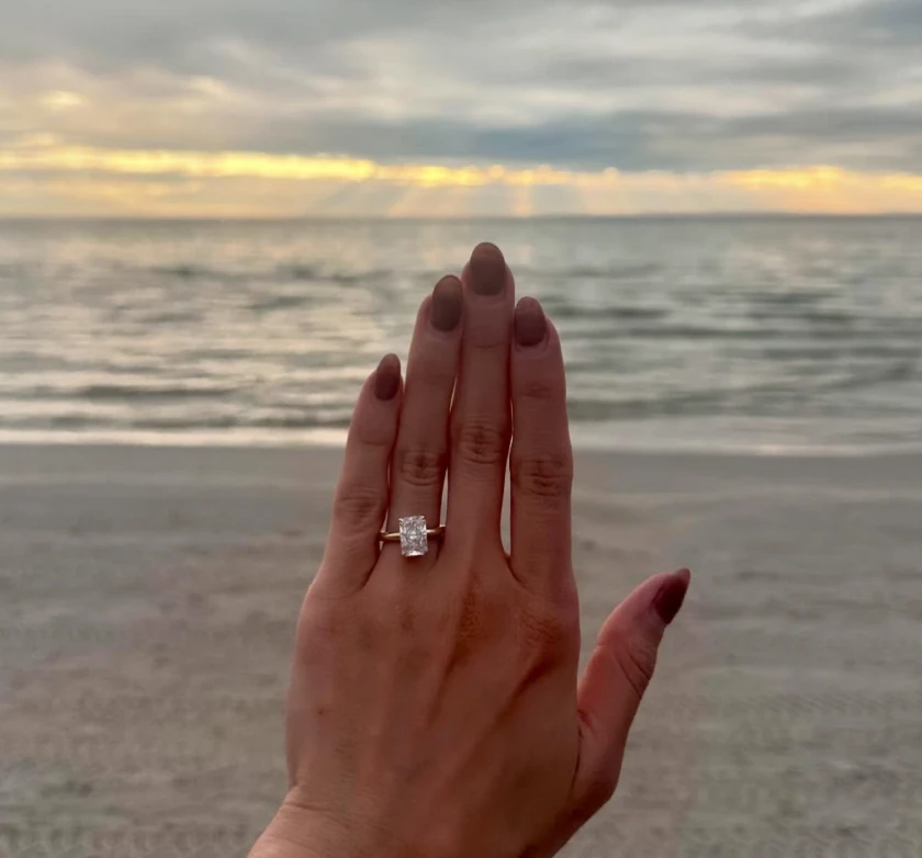 A hand wearing a ring with a rectangular clear centre stone, held in front of the sea.