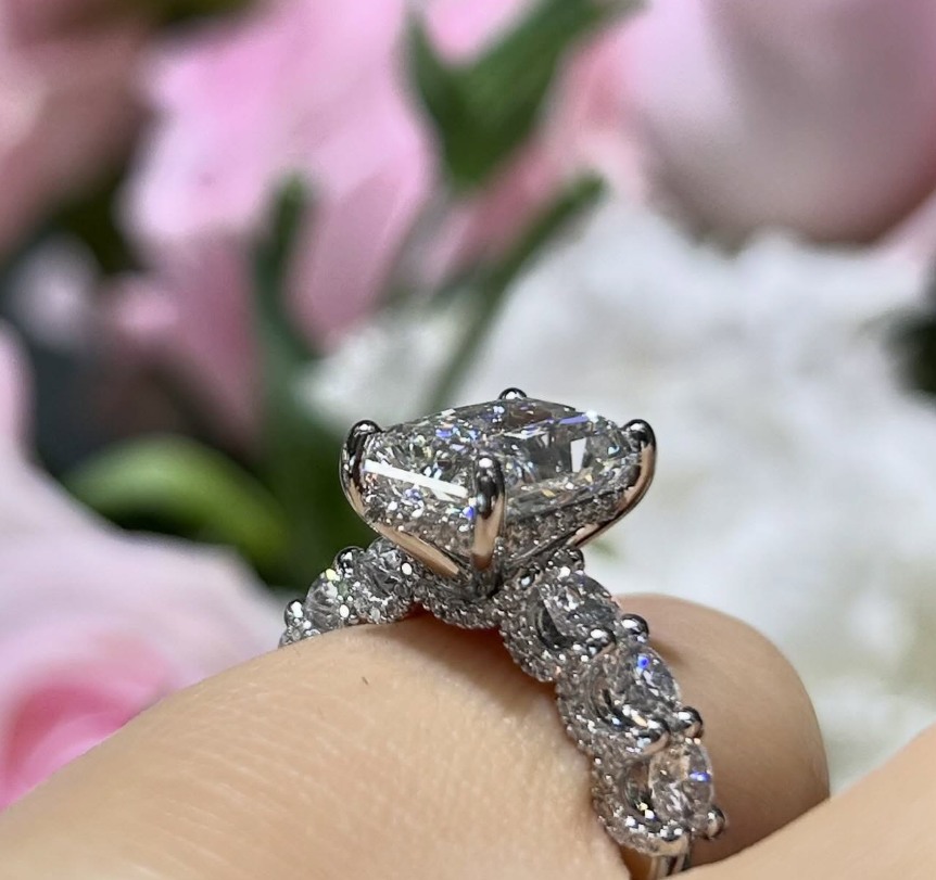 Close side view of a ring on a finger, showing a raised centre setting and large stones along the shoulders, with pink flowers behind.
