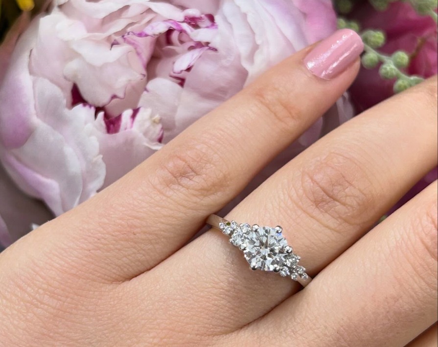 Round centre stone with smaller side stones in an engagement ring worn on a hand beside pink flowers.