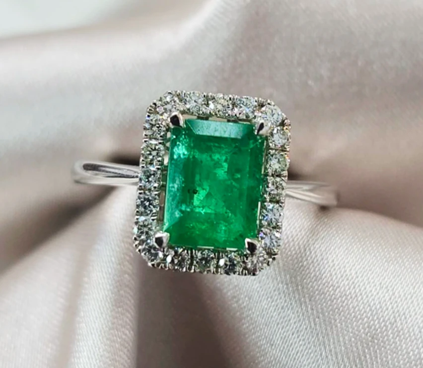 Rectangular green centre stone surrounded by a halo of colourless stones in a ring on pale fabric.