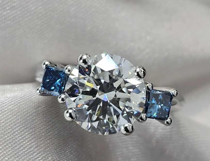 Round colourless centre stone with square blue side stones in a three-stone ring on pale fabric.