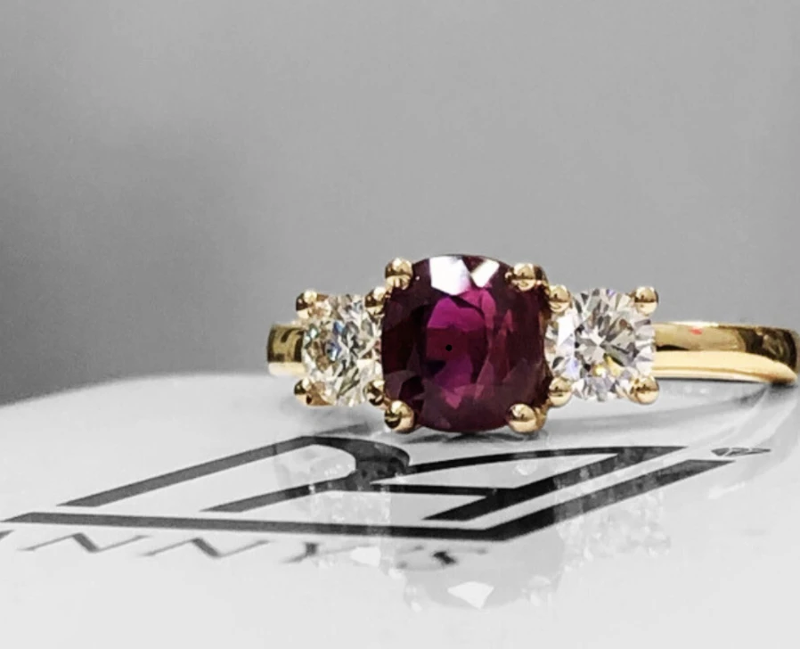 Purplish-red centre ring with two round clear side stones on an Anny’s display card.