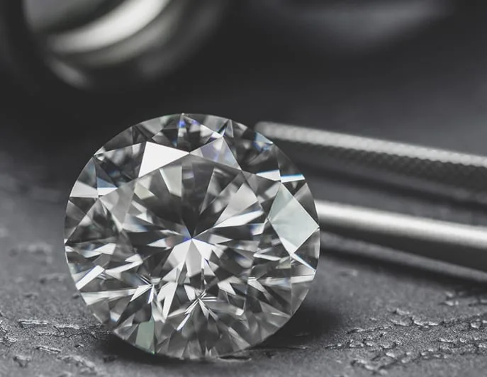 A loose round brilliant diamond on a dark bench beside a pair of jeweller's tweezers