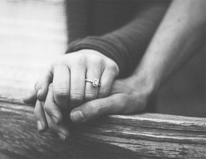 Two hands clasped, one wearing an emerald-cut engagement ring