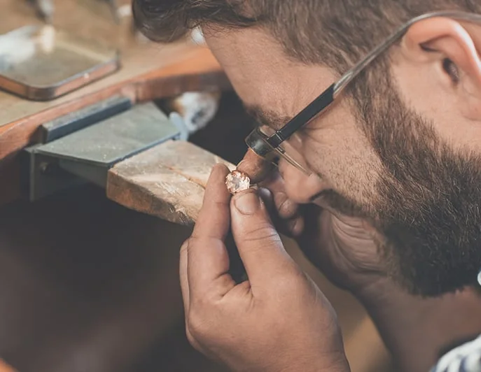 A jeweller holding a loupe to his eye, inspecting the setting of a small pink-stone ring.