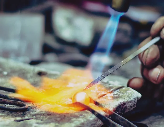 A blue torch flame melting a glowing bead of solder on a soldering block, steadied with a steel pick.