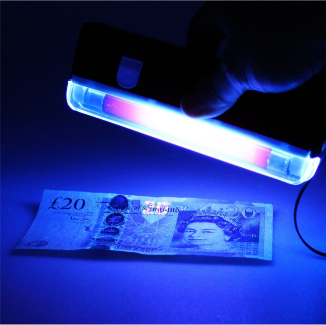A handheld ultraviolet lamp held over a banknote, making its security features glow blue